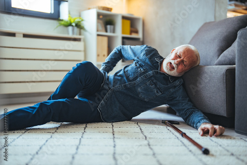 Elderly Senior Man Slip And Fall. Fallen Old Person in the Living Room. Sad tired stylish old man wearing checked shirt making effort trying to get up leaning on cane and divan