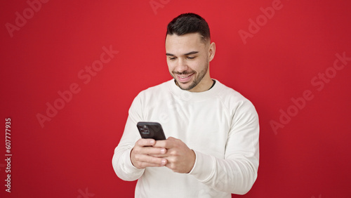 Young hispanic man using smartphone smiling over isolated red background