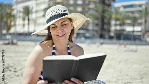Middle age hispanic woman tourist sunbathing sitting on the towel reading a book at the beach