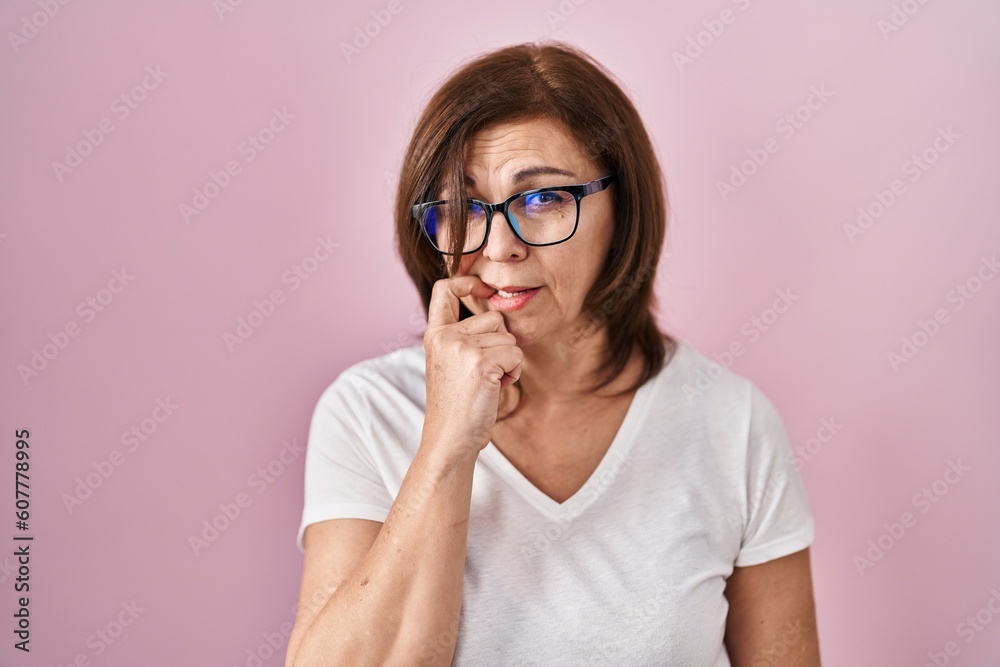Middle age hispanic woman standing over pink background looking stressed and nervous with hands on mouth biting nails. anxiety problem.