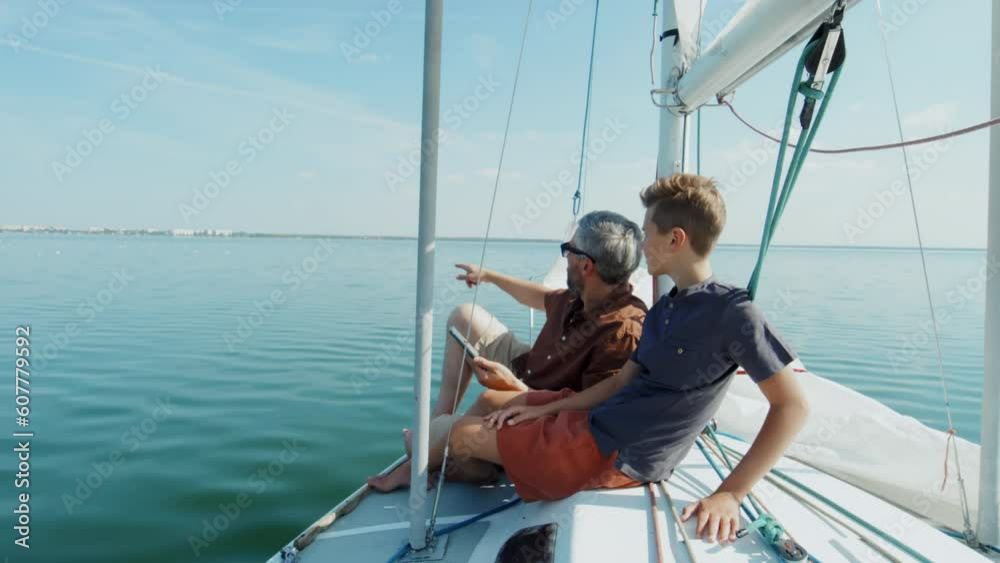 Mature Caucasian man and his teen son sitting relaxed on board the yacht checking direction using map on smartphone Mature Caucasian man and his teen son sitting relaxed on board the yacht checking direction using map on smartphone