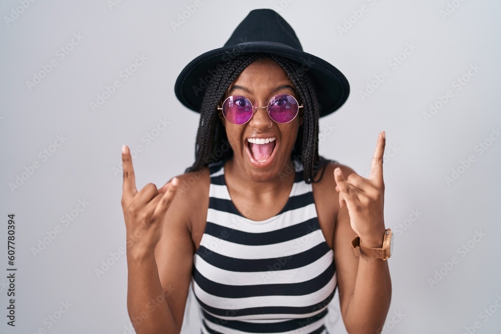 Young african american with braids wearing hat and sunglasses shouting with crazy expression doing rock symbol with hands up. music star. heavy music concept. Young african american with braids wearing hat and sunglasses shouting with crazy expression doing rock symbol with hands up. music star. heavy music concept.