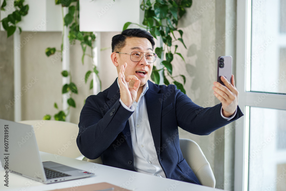 Smiling asian businessman waving hand during video call on smartphone in office.