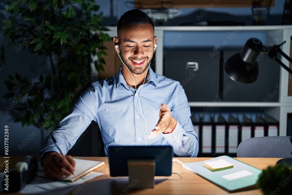 Young hispanic man working at the office at night smiling cheerful offering palm hand giving assistance and acceptance. Young hispanic man working at the office at night smiling cheerful offering palm hand giving assistance and acceptance.