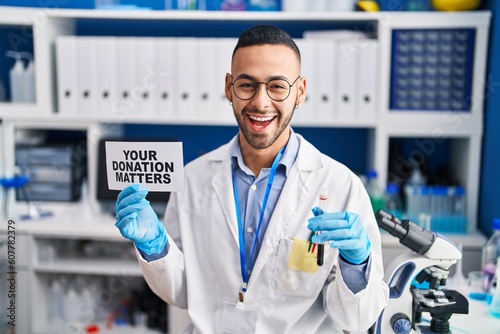 Young hispanic man working at scientist laboratory holding your donation matters holding blood sample celebrating crazy and amazed for success with open eyes screaming excited.