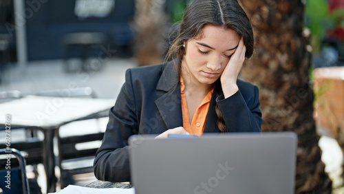 Young beautiful hispanic woman business worker stressed using laptop at coffee shop terrace