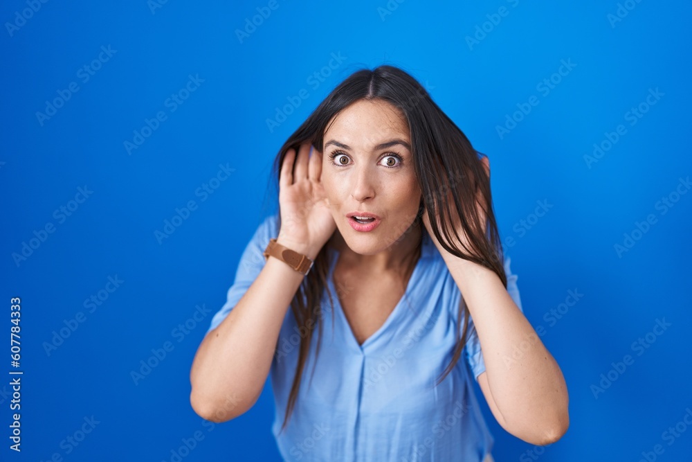 Young brunette woman standing over blue background trying to hear both hands on ear gesture, curious for gossip. hearing problem, deaf Young brunette woman standing over blue background trying to hear both hands on ear gesture, curious for gossip. hearing problem, deaf