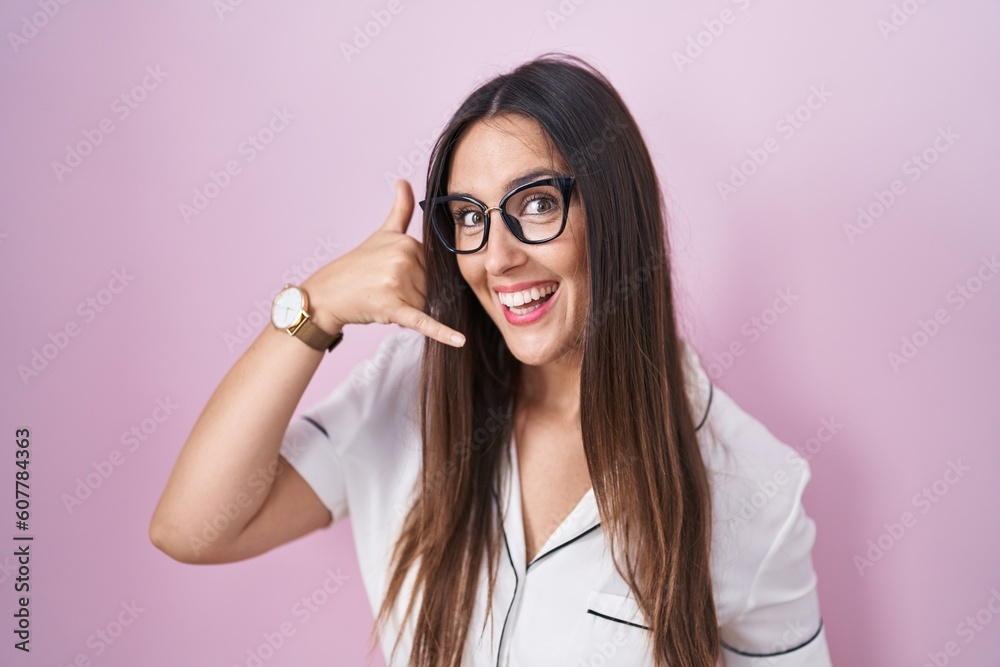 Young brunette woman wearing glasses standing over pink background smiling doing phone gesture with hand and fingers like talking on the telephone. communicating concepts. Young brunette woman wearing glasses standing over pink background smiling doing phone gesture with hand and fingers like talking on the telephone. communicating concepts.