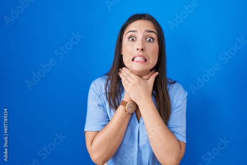 Young brunette woman standing over blue background shouting suffocate because painful strangle. health problem. asphyxiate and suicide concept.
