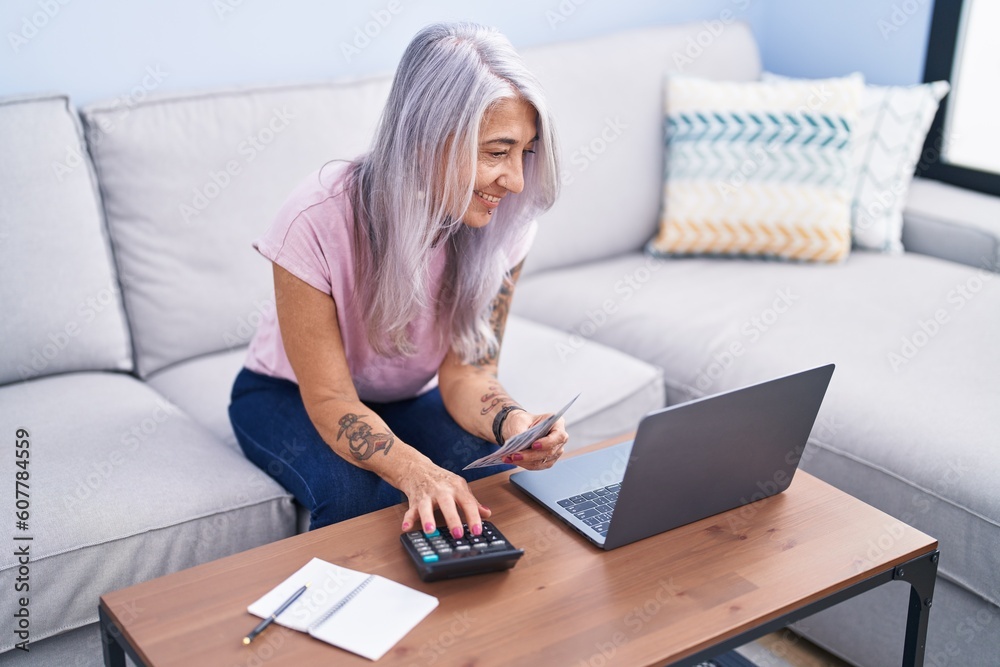 Middle age grey-haired woman using laptop counting dollars at home Middle age grey-haired woman using laptop counting dollars at home