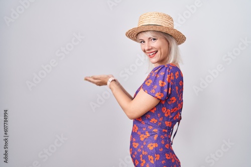 Young caucasian woman wearing flowers dress and summer hat pointing aside with hands open palms showing copy space, presenting advertisement smiling excited happy