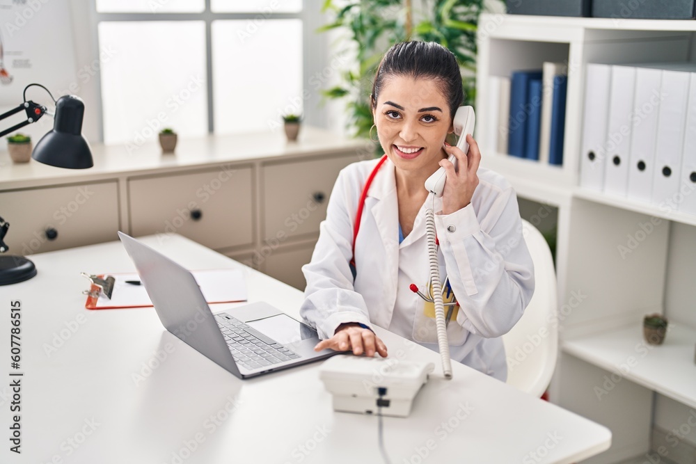 Young beautiful hispanic woman doctor using laptop talking on telephone at clinic Young beautiful hispanic woman doctor using laptop talking on telephone at clinic