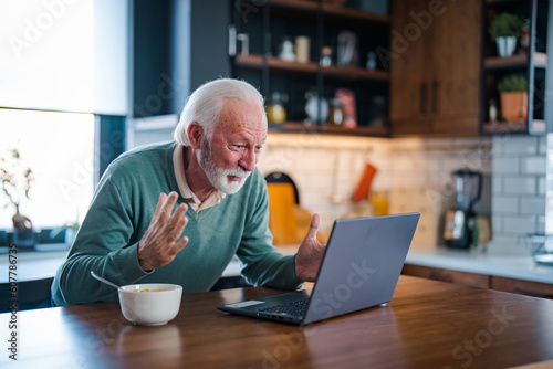 Good-looking enterpreneur working on laptop while having coffee and breakfast. Elderly man using laptop at kitchen table. Computer, money and communication with an elderly male pensioner