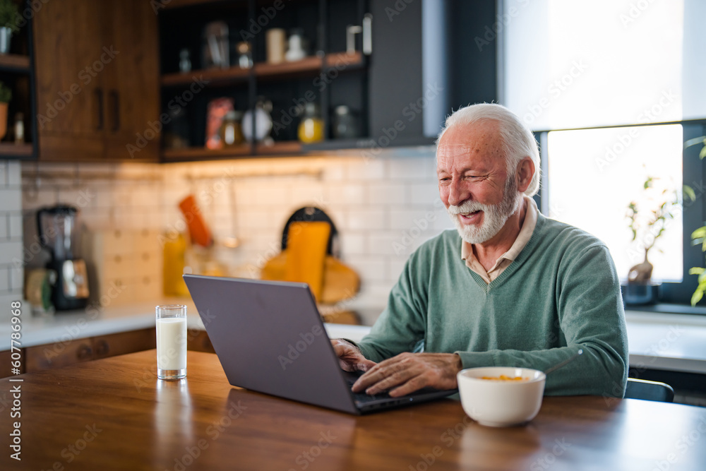 Mature man typing on laptop in kitchen during breakfast and driking coffee. Elderly retired person working from home, telecommuting using remote internet job online communication on modern Mature man typing on laptop in kitchen during breakfast and driking coffee. Elderly retired person working from home, telecommuting using remote internet job online communication on modern