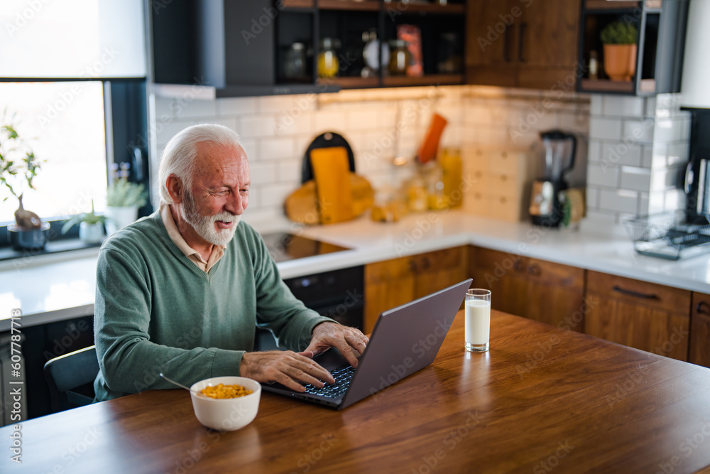 Busy with work. Grey-haired bearded senior man eyeglasses looking concentrated. One senior businessman standing in the kitchen at home and working on the laptop while drinking coffee. Busy with work. Grey-haired bearded senior man eyeglasses looking concentrated. One senior businessman standing in the kitchen at home and working on the laptop while drinking coffee.