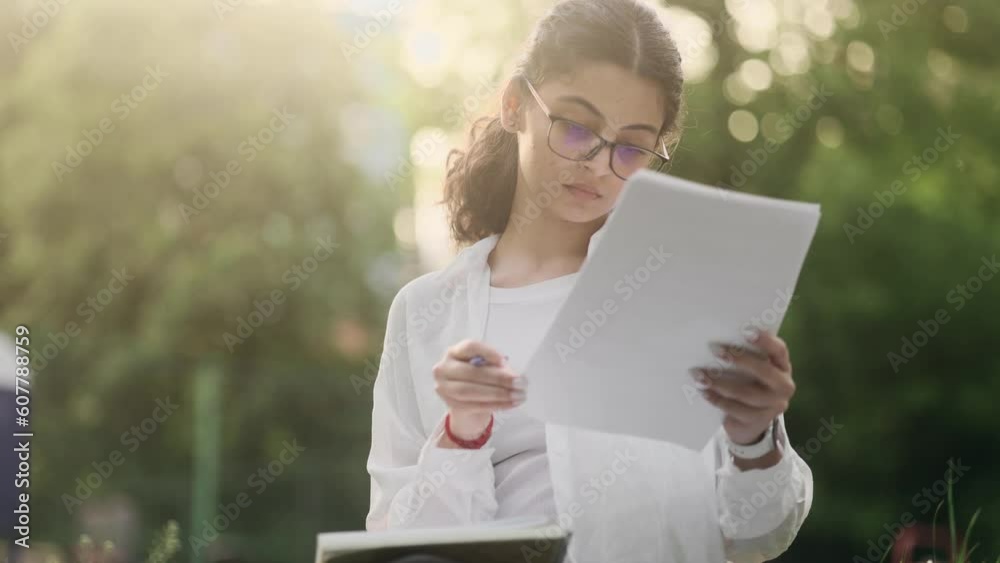 Portrait of focused indian girl with glasses preparing for exam and studying at park Charming student make notes in her notebook while looking at papers outdoors Distance remote education Portrait of focused indian girl with glasses preparing for exam and studying at park Charming student make notes in her notebook while looking at papers outdoors Distance remote education