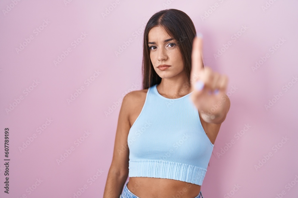 Young brunette woman standing over pink background pointing with finger up and angry expression, showing no gesture