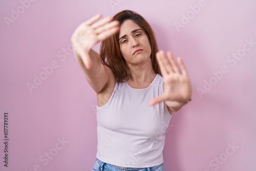 Brunette woman standing over pink background doing frame using hands palms and fingers, camera perspective