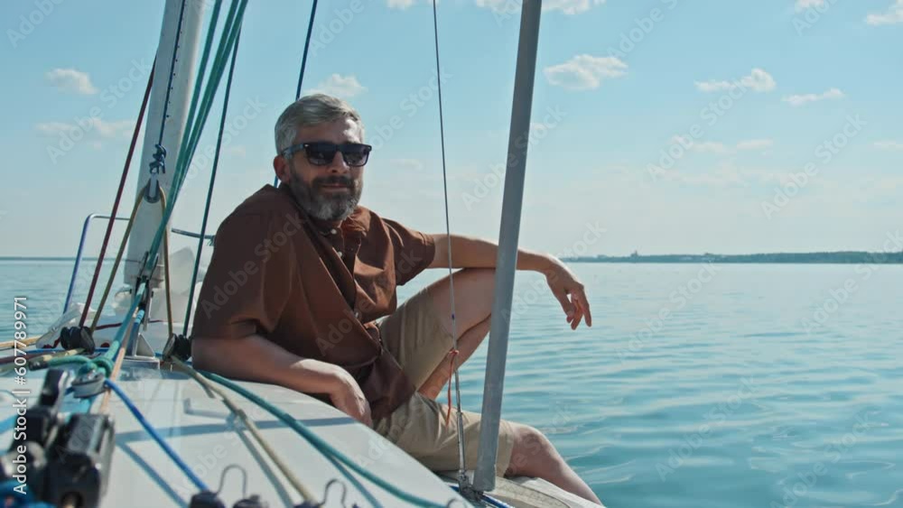 Handsome Caucasian man with beard on face wearing casual clothes with sunglasses sitting relaxed on board the yacht looking at camera Handsome Caucasian man with beard on face wearing casual clothes with sunglasses sitting relaxed on board the yacht looking at camera
