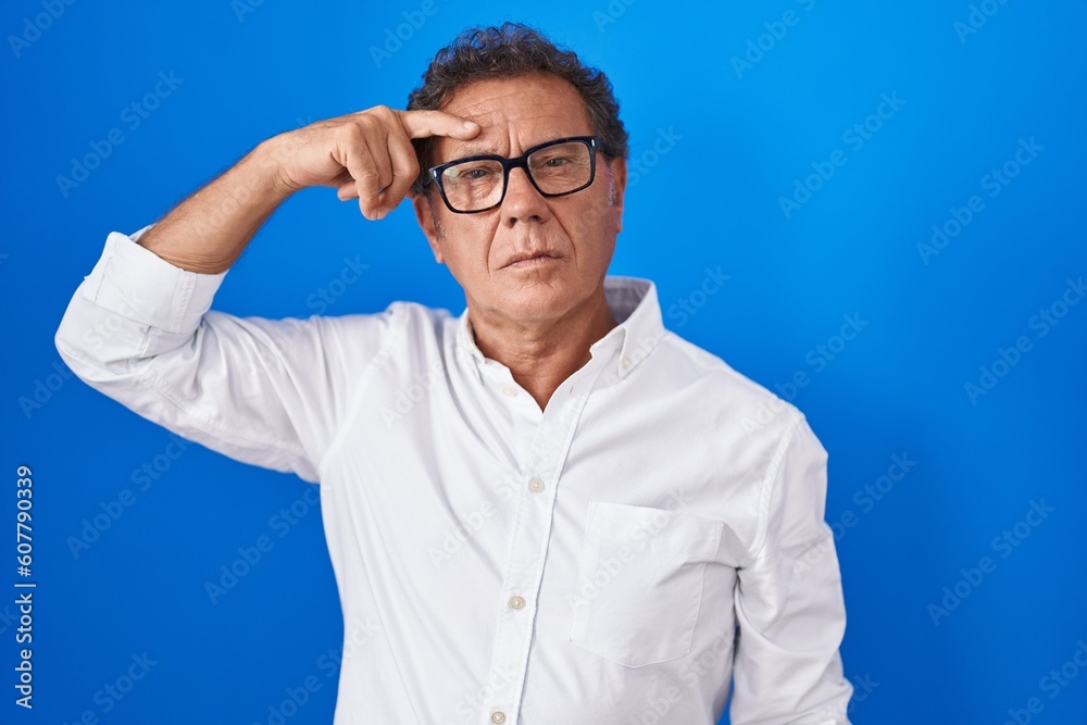 Middle age hispanic man standing over blue background pointing unhappy to pimple on forehead, ugly infection of blackhead. acne and skin problem Middle age hispanic man standing over blue background pointing unhappy to pimple on forehead, ugly infection of blackhead. acne and skin problem