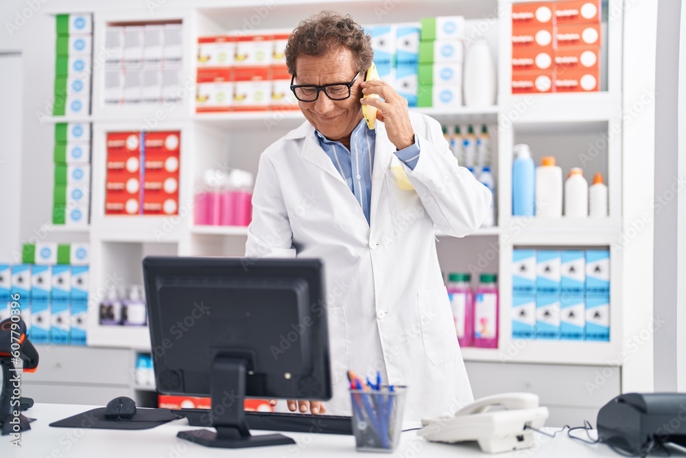 Middle age man pharmacist talking on smartphone using computer at pharmacy