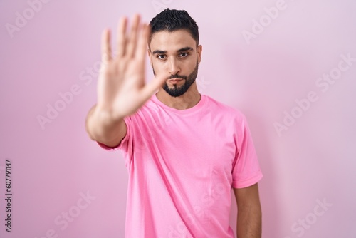 Hispanic young man standing over pink background doing stop sing with palm of the hand. warning expression with negative and serious gesture on the face.