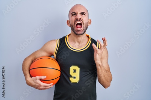 Young bald man with beard wearing basketball uniform holding ball crazy and mad shouting and yelling with aggressive expression and arms raised. frustration concept.