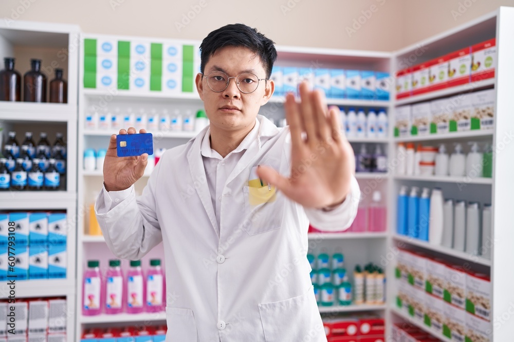 Young asian man working at pharmacy drugstore holding credit card with open hand doing stop sign with serious and confident expression, defense gesture Young asian man working at pharmacy drugstore holding credit card with open hand doing stop sign with serious and confident expression, defense gesture