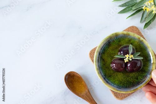 Foto Hand holding bowl with natural extra virgin olive oil and black olives, green tree branch and wooden spoon on white