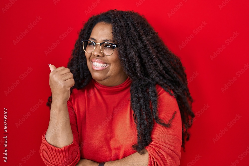 Plus size hispanic woman standing over red background smiling with happy face looking and pointing to the side with thumb up. Plus size hispanic woman standing over red background smiling with happy face looking and pointing to the side with thumb up.