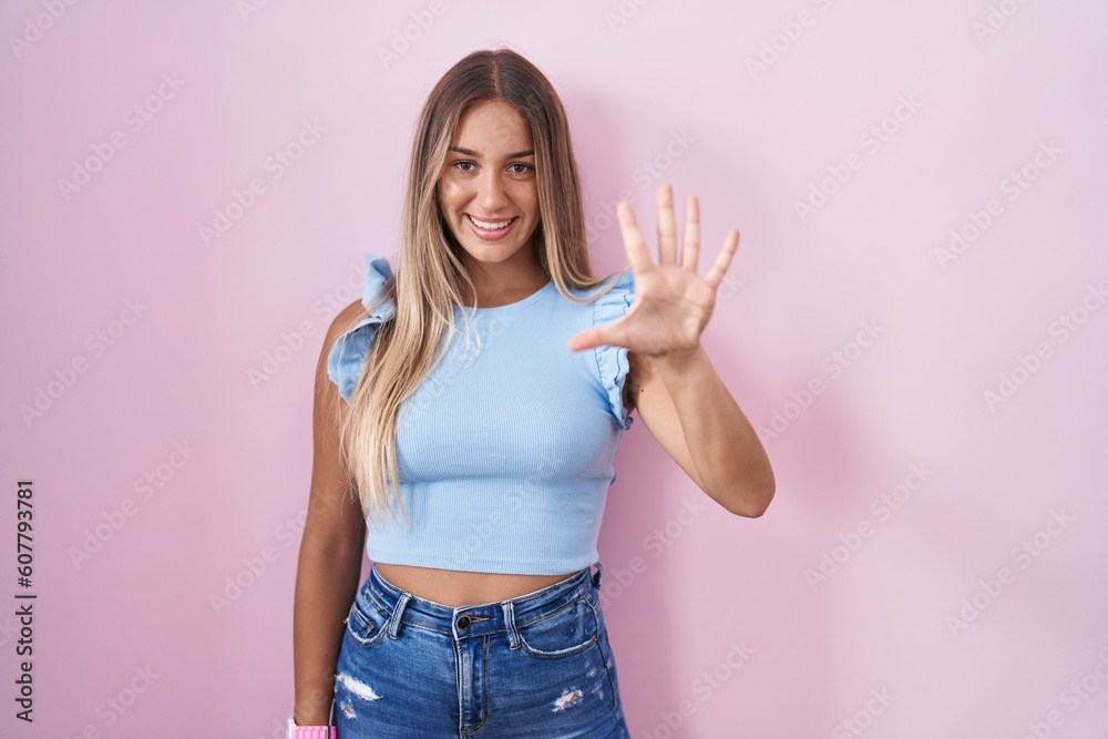Young blonde woman standing over pink background showing and pointing up with fingers number five while smiling confident and happy. Young blonde woman standing over pink background showing and pointing up with fingers number five while smiling confident and happy.