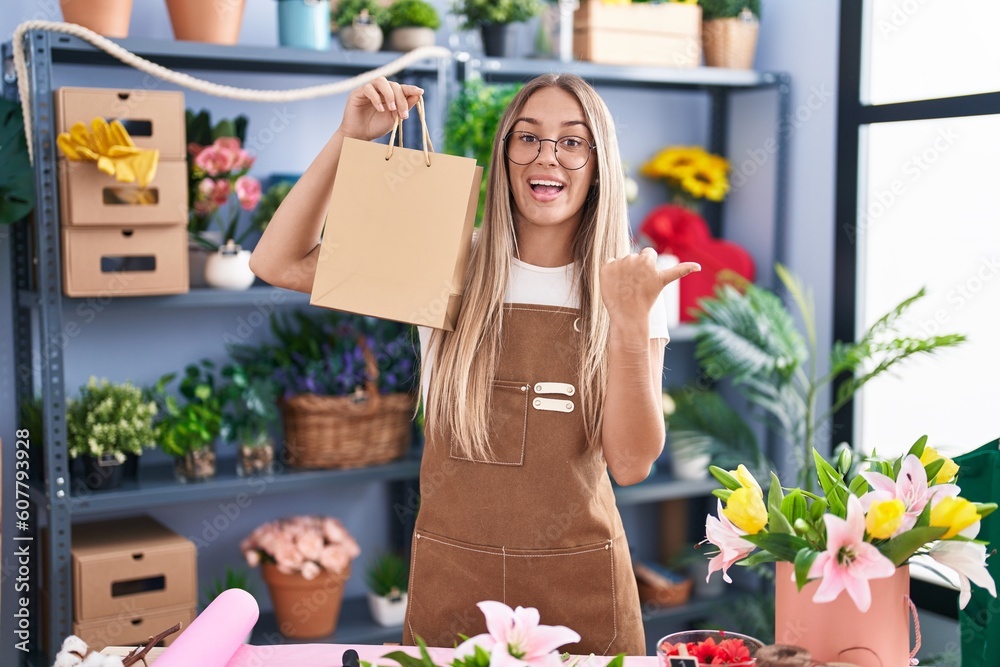 Young blonde woman working at florist shop holding bag pointing thumb up to the side smiling happy with open mouth Young blonde woman working at florist shop holding bag pointing thumb up to the side smiling happy with open mouth