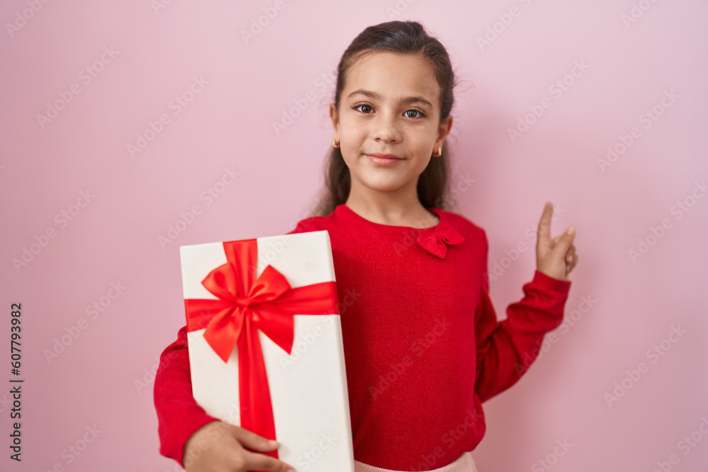 Little hispanic girl holding a gifts smiling happy pointing with hand and finger to the side Little hispanic girl holding a gifts smiling happy pointing with hand and finger to the side