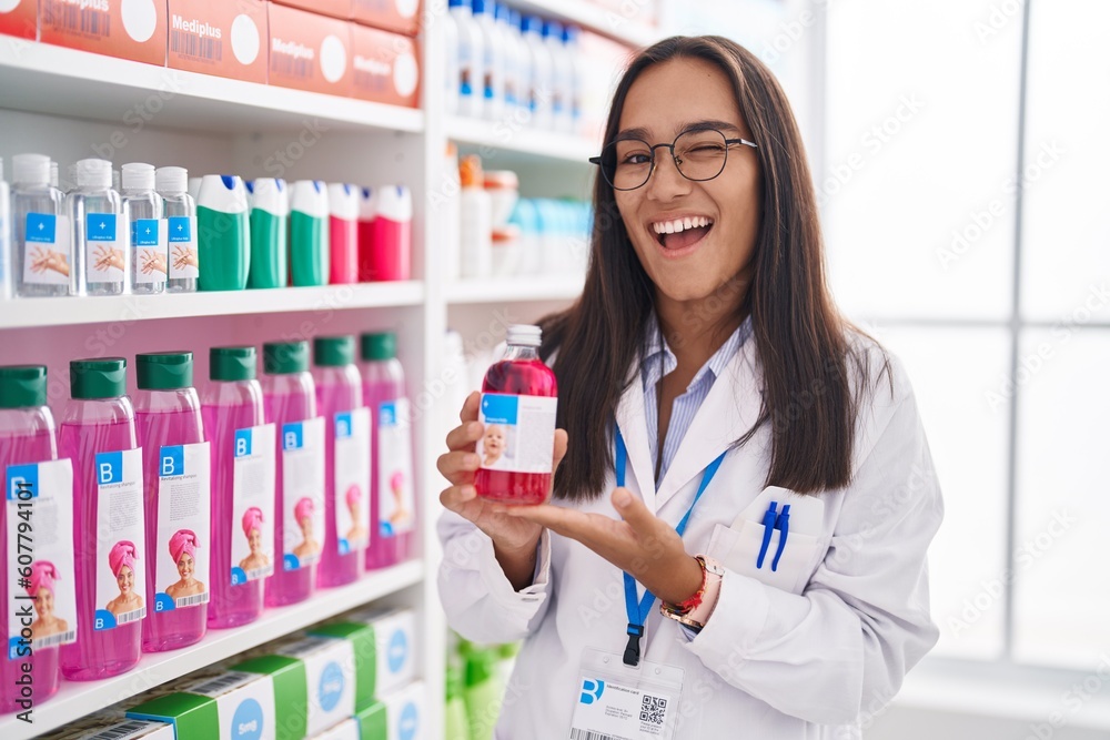 Young hispanic woman working at pharmacy drugstore holding syrup winking looking at the camera with sexy expression, cheerful and happy face.