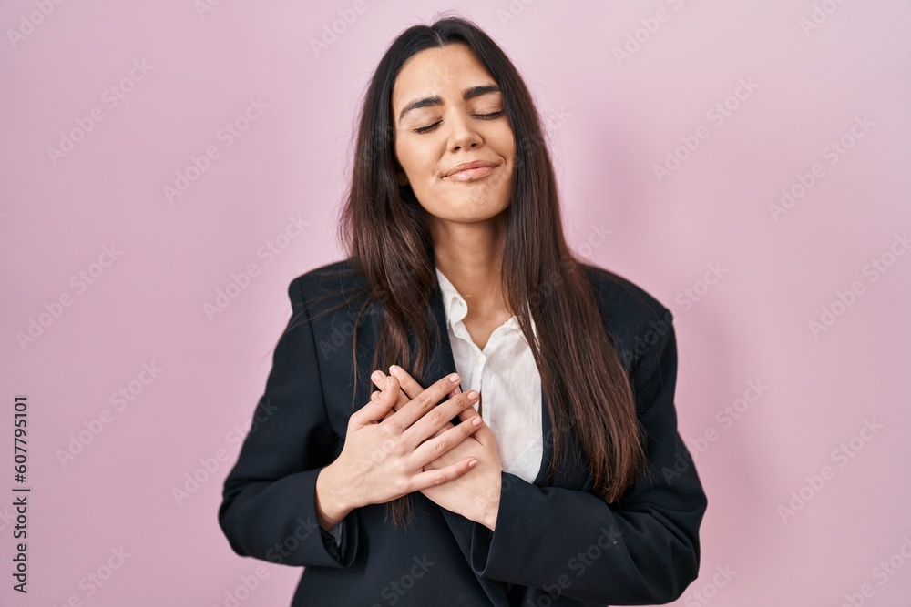 Young brunette woman wearing business style over pink background smiling with hands on chest with closed eyes and grateful gesture on face. health concept. Young brunette woman wearing business style over pink background smiling with hands on chest with closed eyes and grateful gesture on face. health concept.