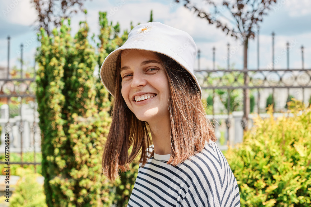 Portrait of young caucasian woman girl with toothy smile walking in green street outdoors looking at camera wearing in striped shirt and panama enjoying walking Portrait of young caucasian woman girl with toothy smile walking in green street outdoors looking at camera wearing in striped shirt and panama enjoying walking