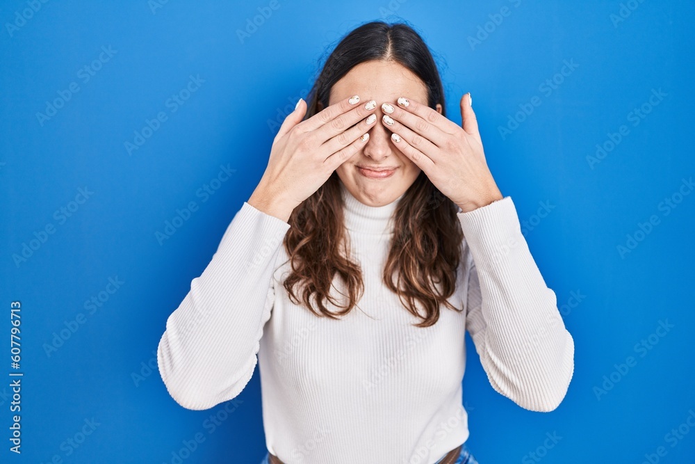 Young hispanic woman standing over blue background covering eyes with hands smiling cheerful and funny. blind concept. Young hispanic woman standing over blue background covering eyes with hands smiling cheerful and funny. blind concept.