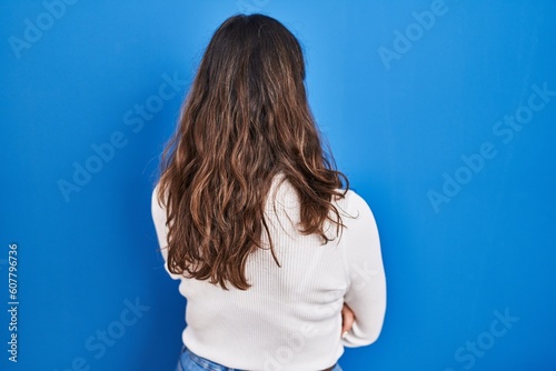 Young hispanic woman standing over blue background standing backwards looking away with crossed arms