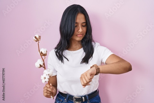 Brunette woman standing over pink background checking the time on wrist watch, relaxed and confident