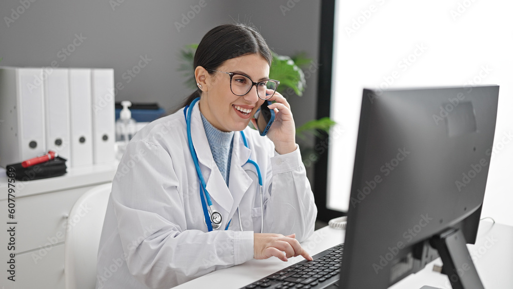Young hispanic woman doctor using computer speaking on smartphone at clinic Young hispanic woman doctor using computer speaking on smartphone at clinic
