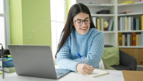Young hispanic woman student using computer taking notes at library university