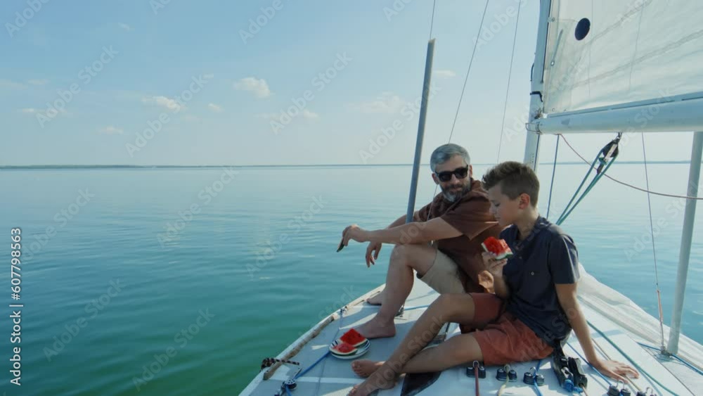 Mature Caucasian man and his teen son spending summer day together on sailboat eating watermelon Mature Caucasian man and his teen son spending summer day together on sailboat eating watermelon
