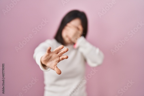 Woman with down syndrome standing over pink background covering eyes with hands and doing stop gesture with sad and fear expression. embarrassed and negative concept.