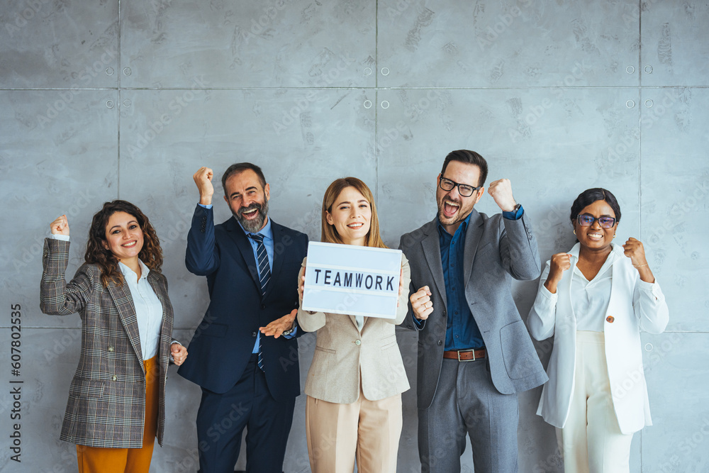 Fototapeta premium Portrait of multi ethnic successful business team looking at camera in the office and team leader holding sign ''teamwork'' in hands. Business men and women managers team holding big teamwork banner.