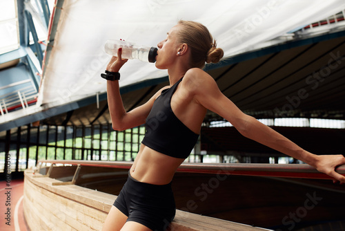 A young girl athlete drinking water from a sports bottle is resting after training on the track. Slender athletic blonde in a black tracksuit in a relaxation pose.