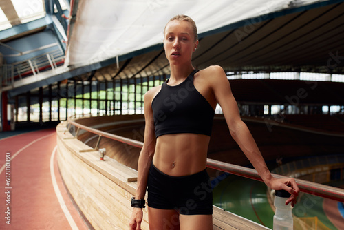 A young girl athlete is resting after a workout on the track. Slender athletic blonde in a black tracksuit in a relaxation pose. Fitness and relaxation concept.