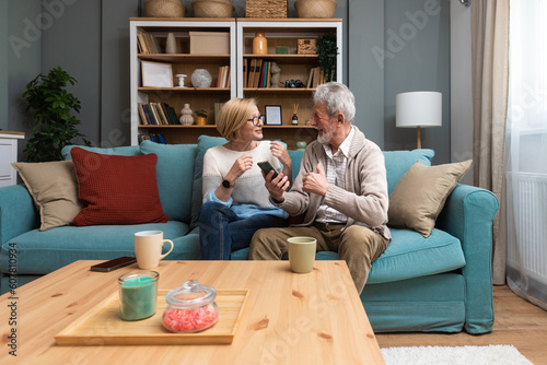 Relaxed mature 60s woman and 70s man older aged senior couple customer holding smartphone using mobile app texting message search ecommerce offers on cellphone technology device sits on sofa at home