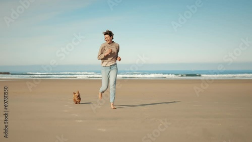 Small brown dog and blonde woman running in slow motion at the seaside on a sunny summer day. Little Havanese puppy and his owner enjoying day at the beach by the blue sea.