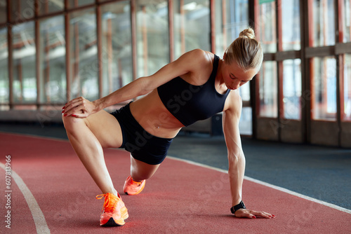Young athlete girl is warming up before the race at the stadium. A slender blonde in a black tracksuit stretches her leg muscles before training. Sports and recreation.