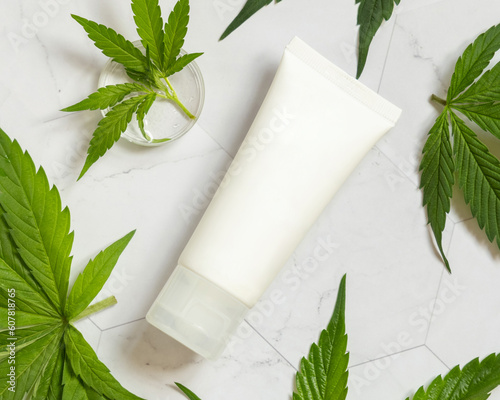 Blank Cream tube near green cannabis leaves on white table, top view. Cosmetic Mockup